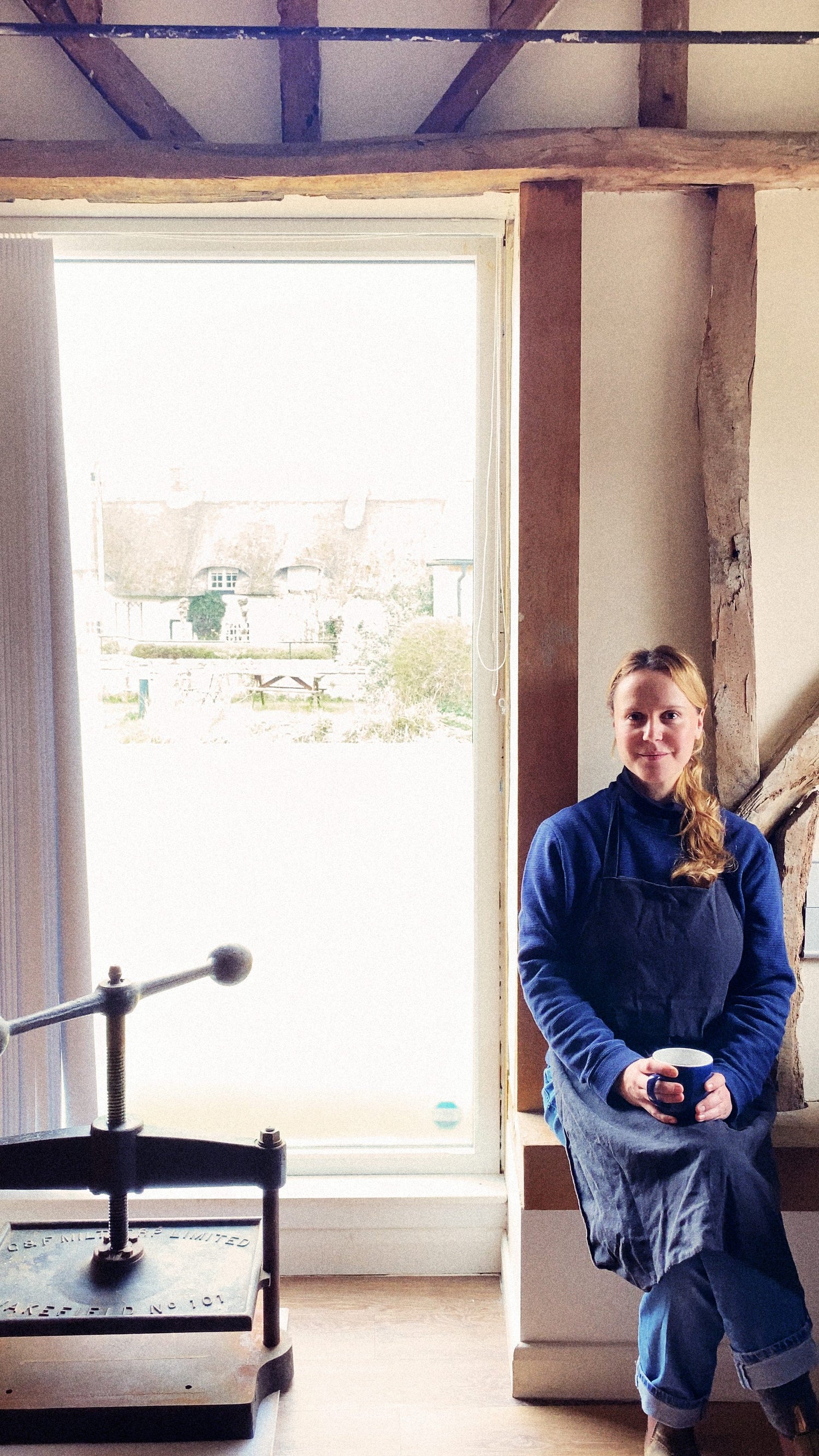 Metta's founder, Kathrin, at a window, a mug of tea in hand — weathered timber beams framing the space.