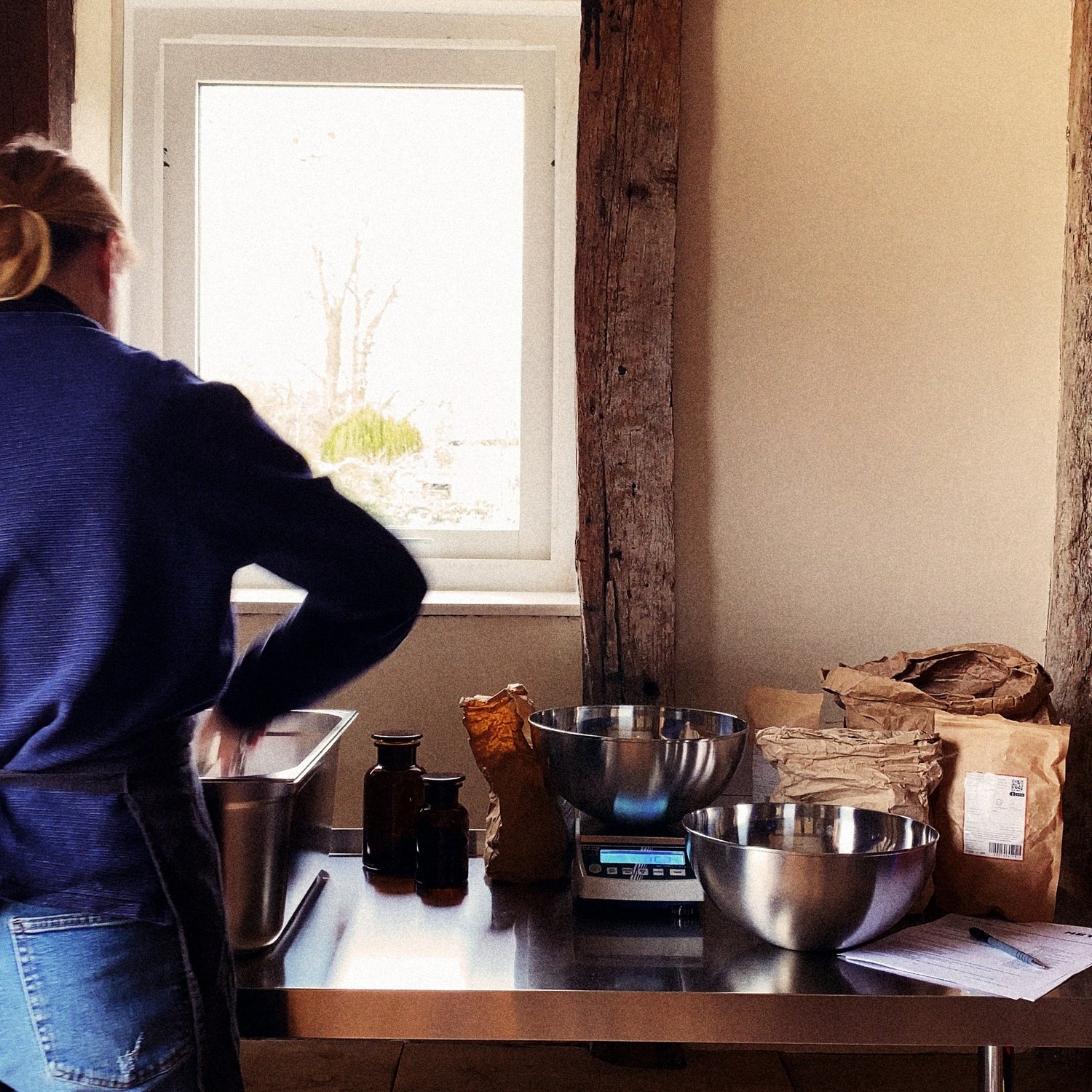 Metta's founder blending herbs — stainless-steel table, mixing bowls, brown apothecary bottles and sacks of dried herbs.