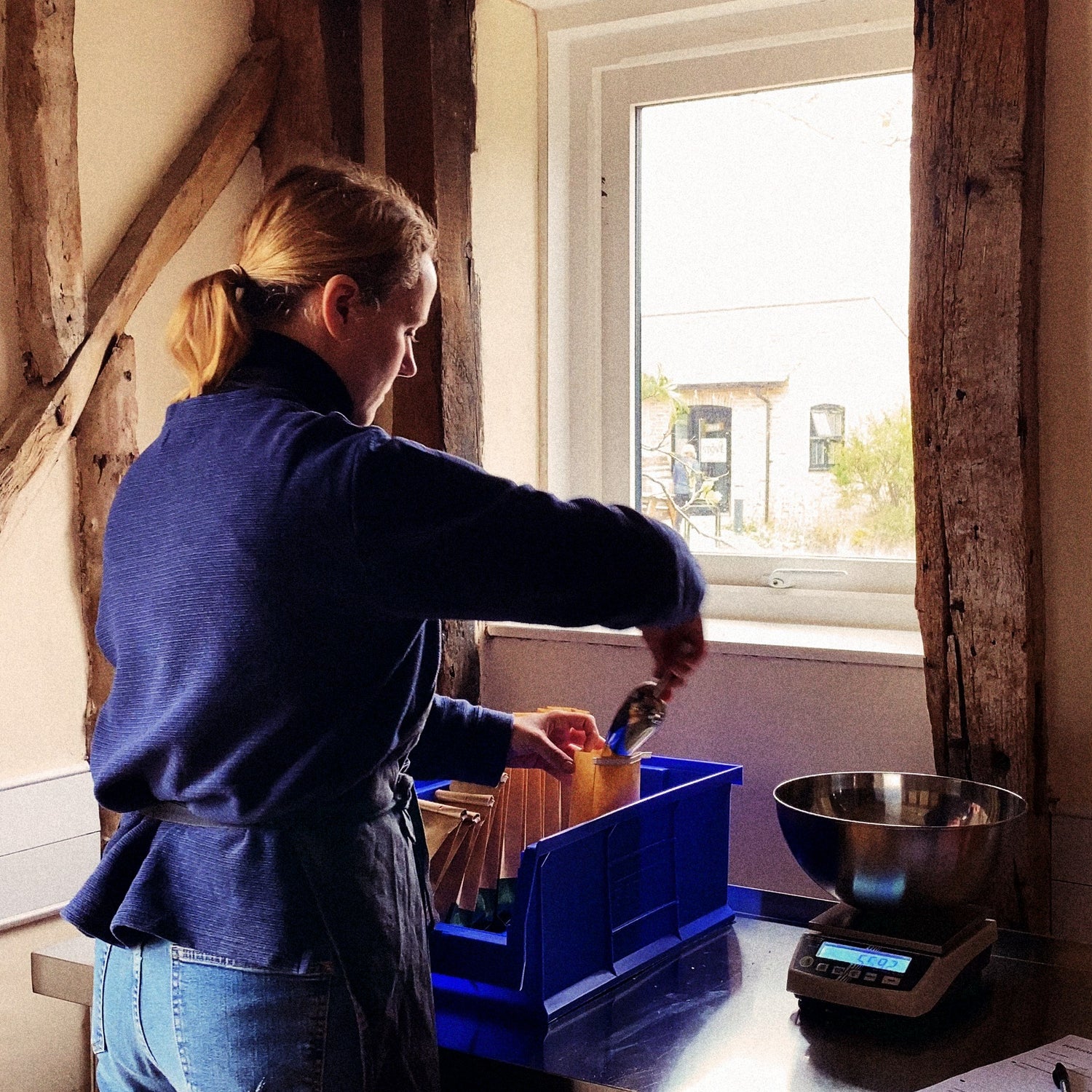 Metta's founder scooping dried herbs from a small crate at the blending bench, weighing each portion on a steel scale.