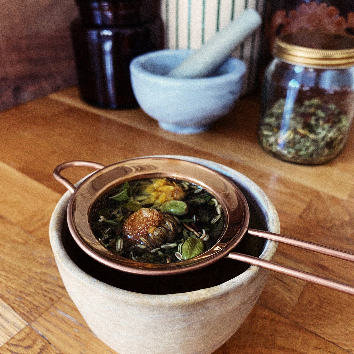 A copper tea strainer resting on a stoneware cup — spent herbs from a freshly brewed blend.