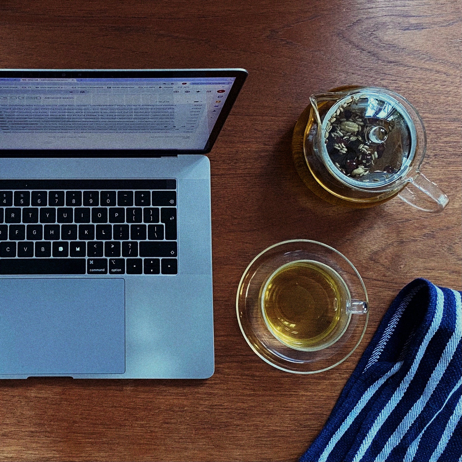 An open laptop on a wooden desk — a glass teapot of brewing herbs and a folded tea towel alongside.